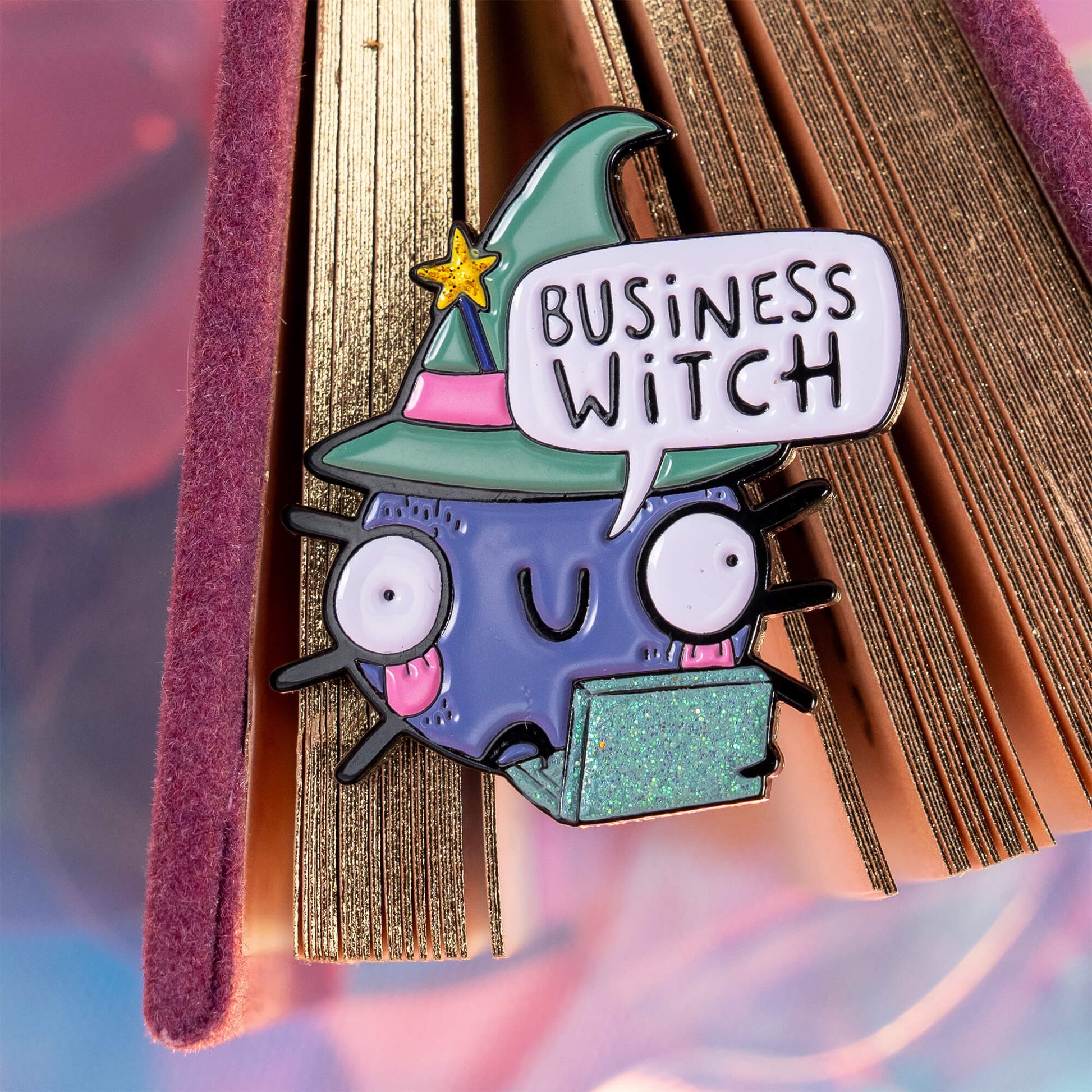 An enamel pin on the edge of a book with gold-edged pages. The pin displays a cute, wide-eyed cartoon character in a green witch hat, holding a sparkly green laptop, with a speech bubble saying 'Business Witch'. The background is a colourful mix of pastel pinks and blues. Brand: Happiness Enchanters.