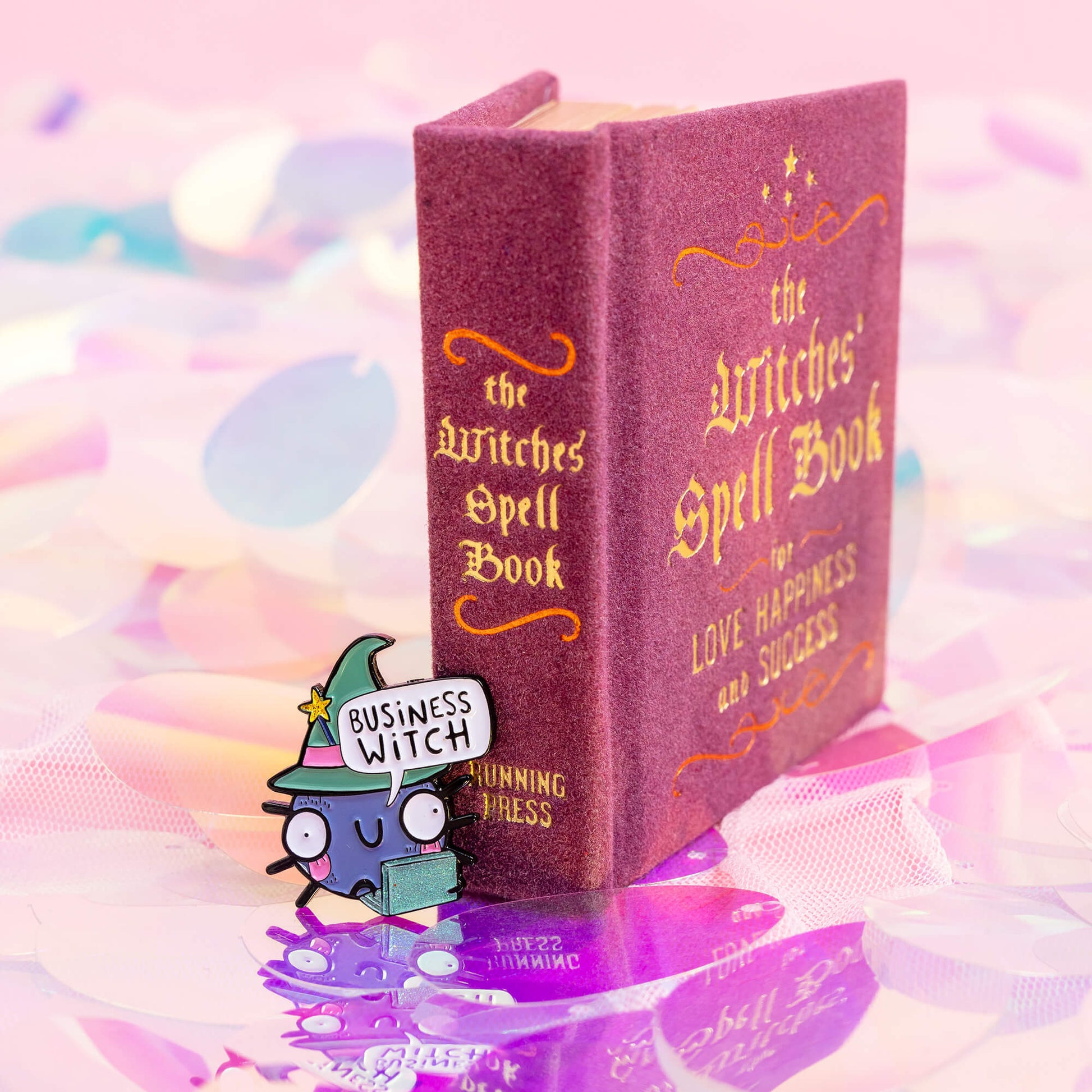 An enamel pin featuring a cute, wide-eyed cartoon character with a speech bubble that says 'Business Witch'. The character is wearing a green witch hat with a yellow star and holding a sparkly green laptop. The background is a colourful, abstract design with pink and blue hues. Brand: Happiness Enchanters.