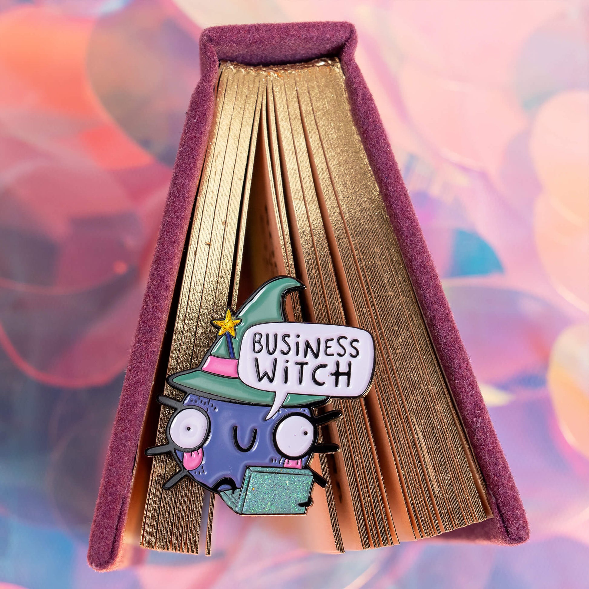 Close-up of an enamel pin attached to the spine of a book with gold-edged pages. The pin shows a cute, wide-eyed cartoon character wearing a green witch hat and holding a sparkly green laptop. A speech bubble above the character reads 'Business Witch'. The book and the background have a whimsical, pastel-coloured design. Brand: Happiness Enchanters.