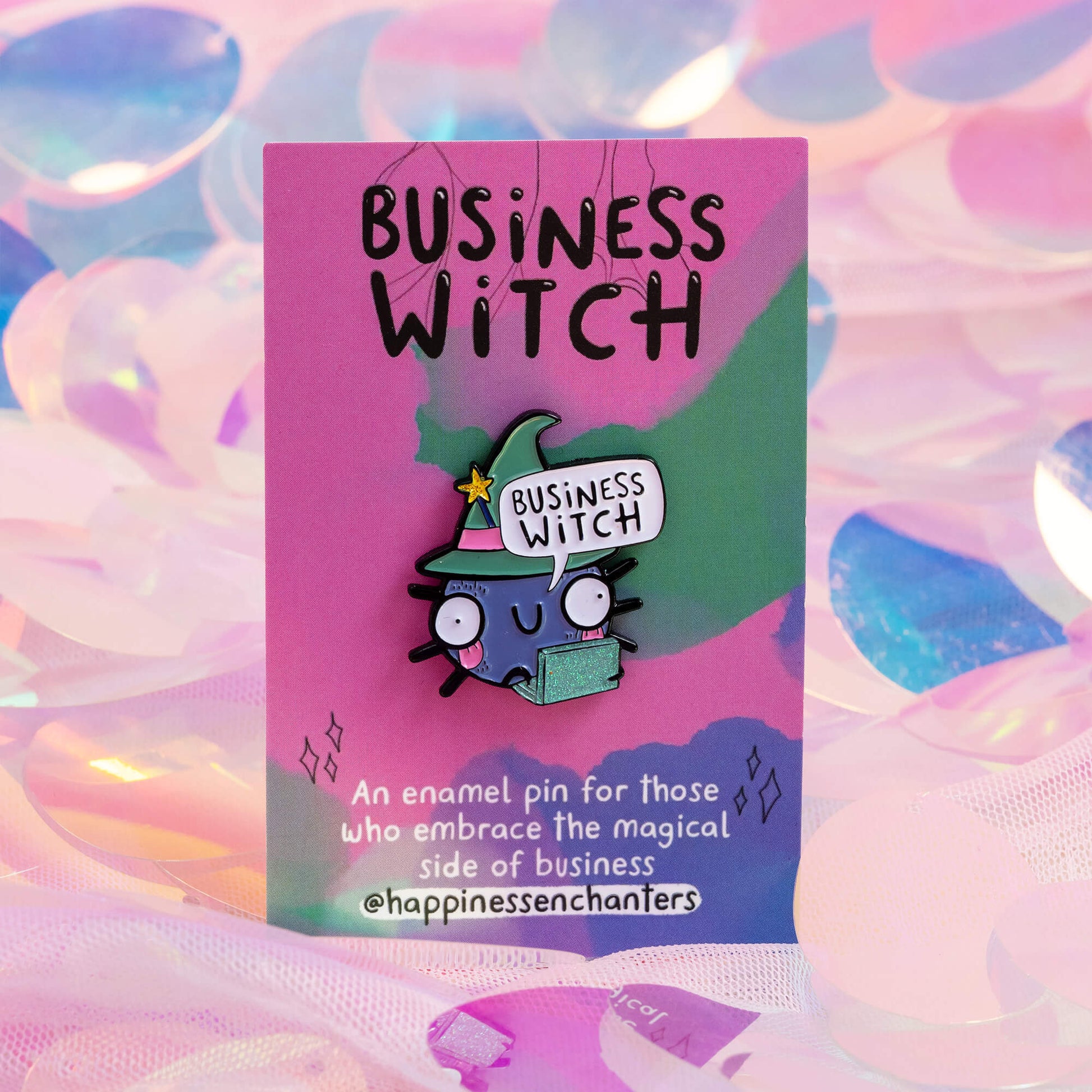 An enamel pin mounted on a pink and green card that reads 'Business Witch' at the top. The pin features a cute, wide-eyed cartoon character wearing a green witch hat with a yellow star and holding a sparkly green laptop. The card has a whimsical, colourful background. Brand: Happiness Enchanters.