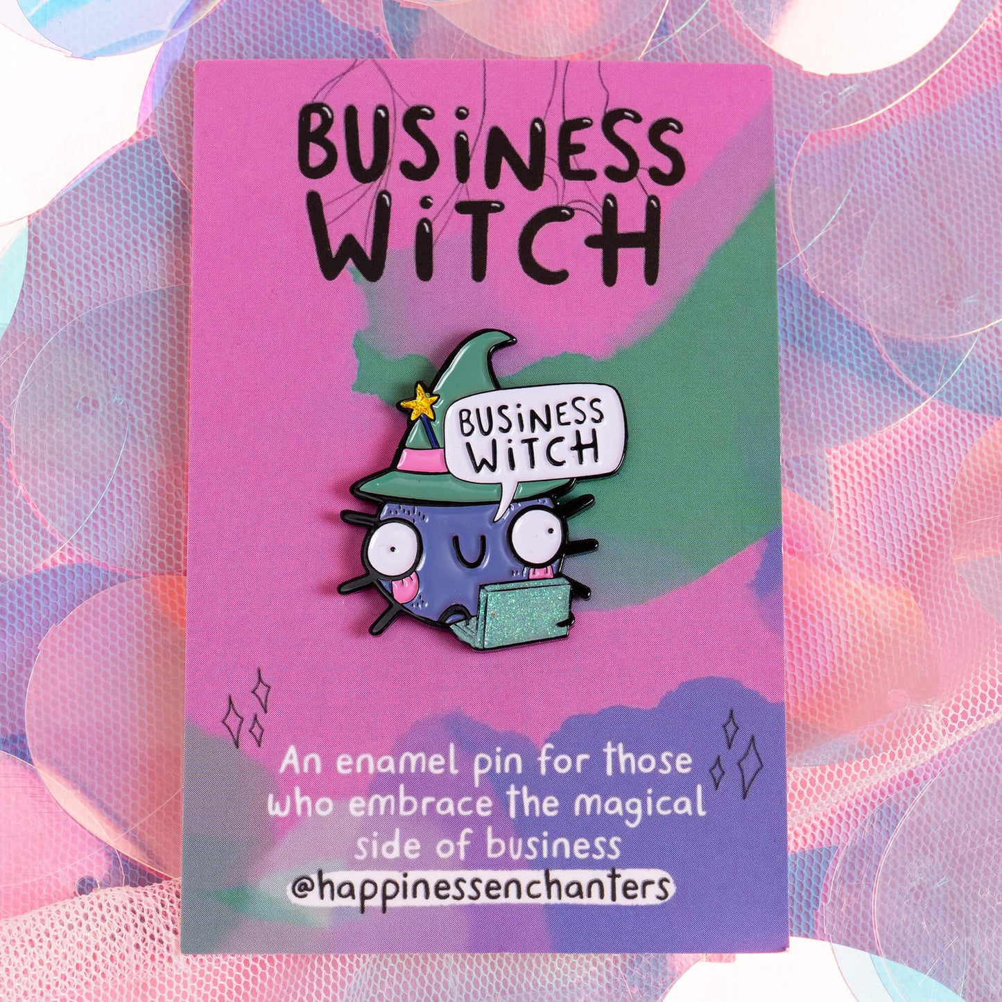 An enamel pin attached to a pink and green card that reads 'Business Witch' at the top. The pin shows a cute, wide-eyed cartoon character wearing a green witch hat with a yellow star, holding a sparkly green laptop, with a speech bubble saying 'Business Witch'. The background is a colourful, pastel design. Brand: Happiness Enchanters.