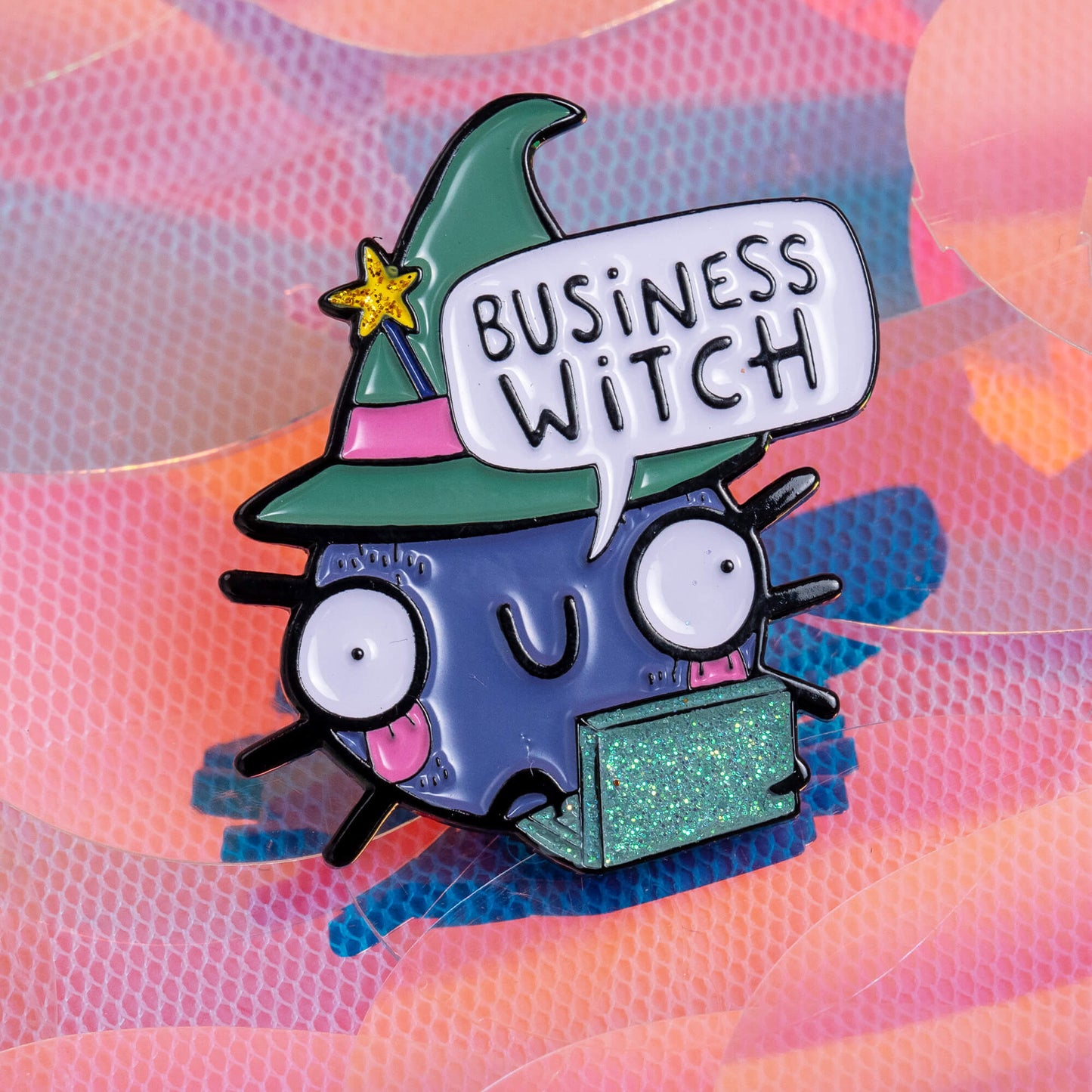 An enamel pin displayed next to a purple book titled 'The Witches' Spell Book for Love, Happiness, and Success'. The pin features a cute, wide-eyed cartoon character in a green witch hat, holding a sparkly green laptop, with a speech bubble that reads 'Business Witch'. The background is a pastel, colourful mix of pinks, purples, and blues. Brand: Happiness Enchanters.