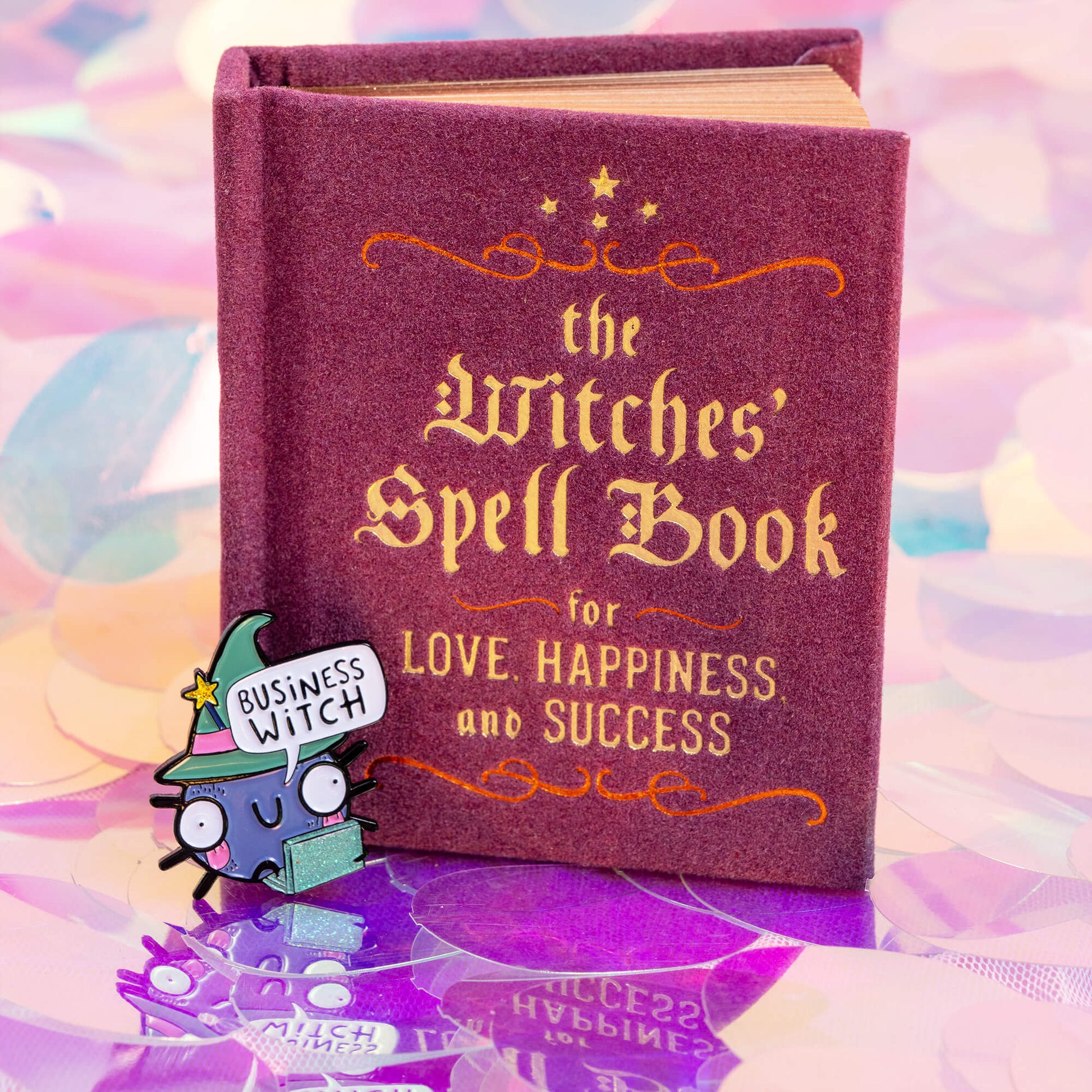 An enamel pin displayed next to a book titled 'The Witches' Spell Book for Love, Happiness, and Success'. The pin features a cute, wide-eyed cartoon character in a green witch hat, holding a sparkly green laptop, with a speech bubble that says 'Business Witch'. The background is a pastel, colourful mix of pinks, purples, and blues. Brand: Happiness Enchanters.