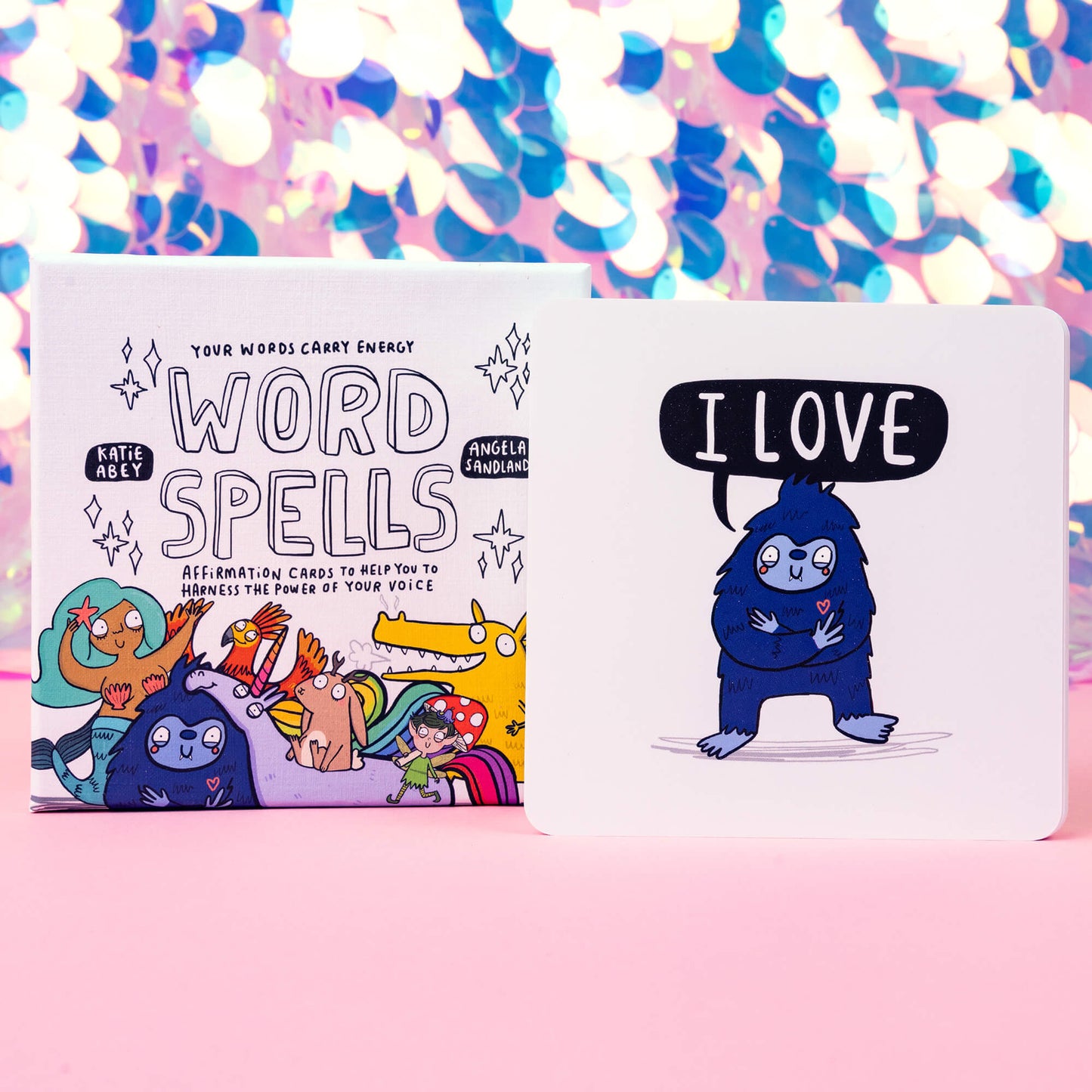 Box of Word Spell Cards on a pink and sparkly background, the box is white with 'your words carry energy' and 'affirmation cards to help you to harness the power of your voice' written on with various illustrations.