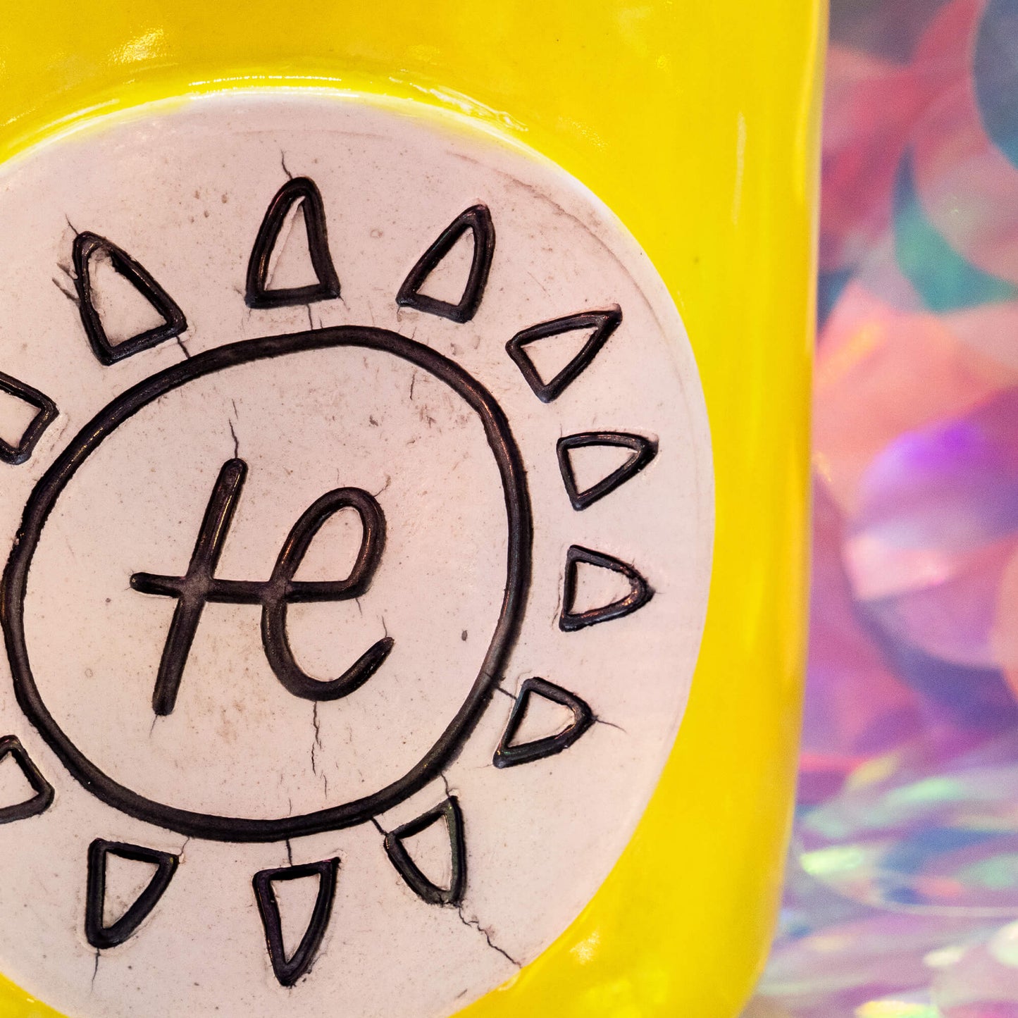 Bright yellow handmade ceramic mug with the Happiness Enchanters sun and symbol design on a white medallion, featuring a glossy pink interior, photographed against a pastel iridescent background.